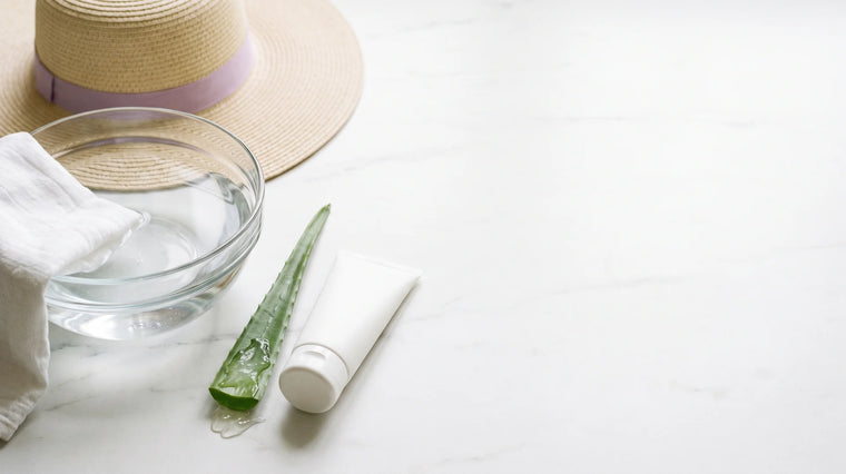 Mermer tezgâhta serin su kâsesi ve bez, aloe vera yaprağı, güneş kremi tüpü ve hasır şapka